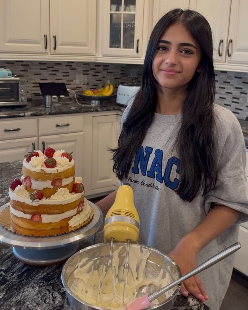 Elina beside a two-tier naked cake topped with strawberries and cream, with a hand mixer in a bowl of whipped frosting