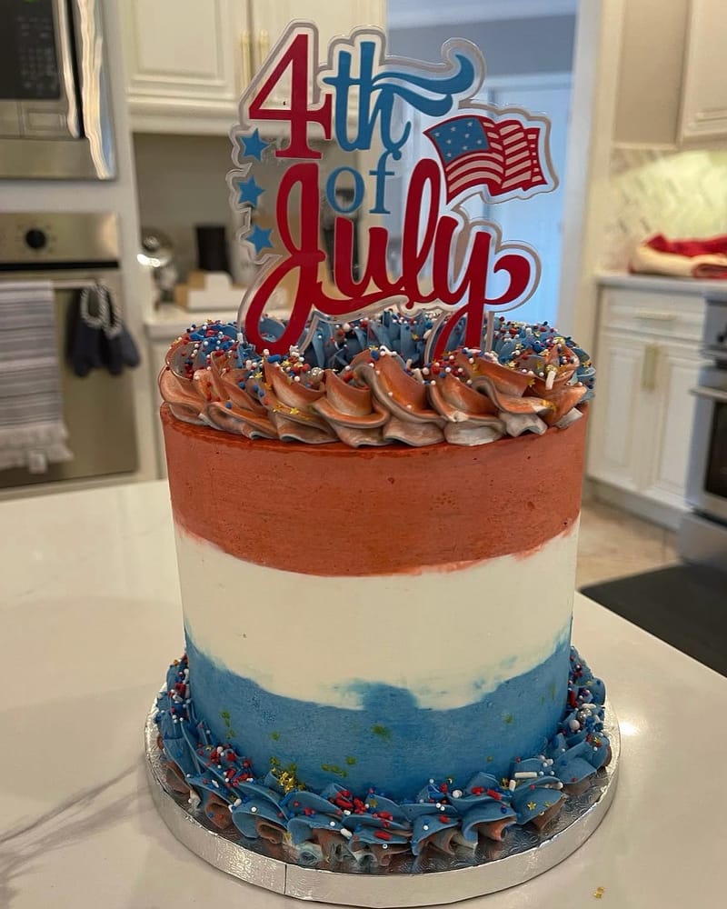 Tall Fourth of July cake frosted in red, white and blue bands, with piped swirls, sprinkles and a 4th of July topper