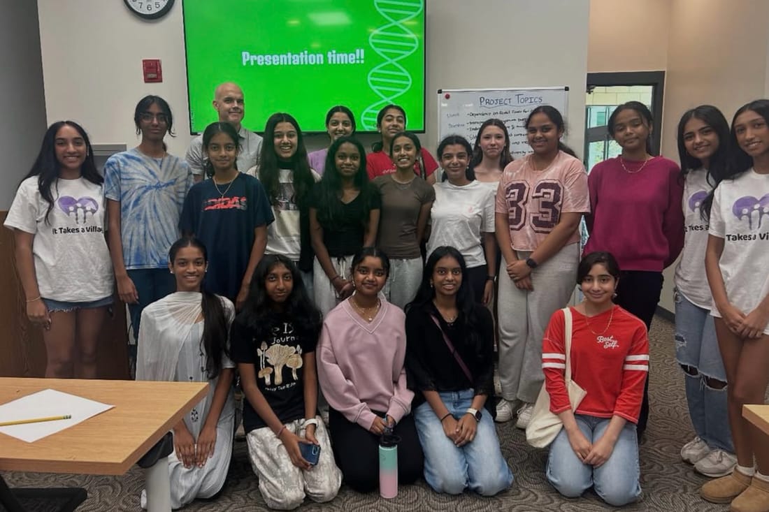 About twenty students, several in It Takes A Village T-shirts, in front of a Presentation time slide and a whiteboard of project topics
